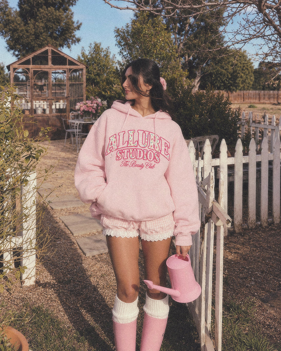 The Beauty Club Hoodie in dusty pink by a garden gate