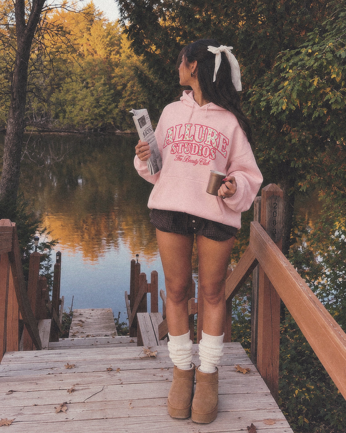 The Beauty Club Hoodie in dusty pink on a lake dock